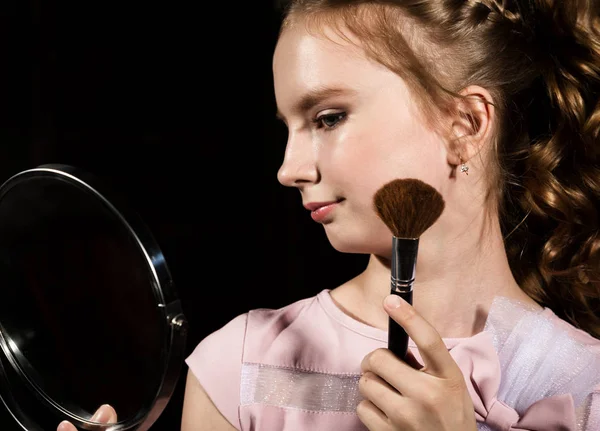 Маленькая девочка с круглым зеркалом делает макияж на темном фоне — стоковое фото