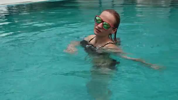 Belle jeune femme heureuse en lunettes de soleil souriante et relaxante dans la piscine de l'hôtel — Video