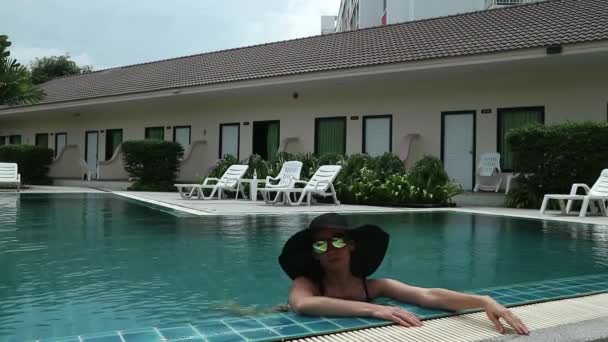 Молодая женщина в черных широких краях и солнечных очках отдыхает в бассейне — стоковое видео