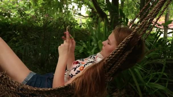 Girl relaxing in hammock uses a smartphone, swaying gently near hotel on beach — Stock Video