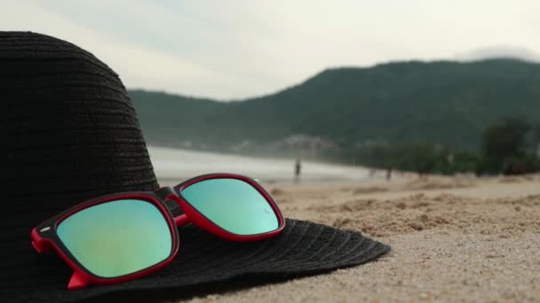 Gafas de sol y ala ancha negra tumbadas sobre una arena. En unas gafas de sol reflejadas mar, olas y cielo . — Vídeo de stock