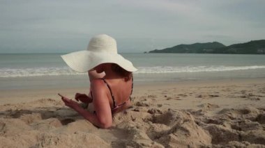 Kumsalda güneşlenme sırasında cep telefonu kullanan genç kadın. Bikini ve güneş şapkalı güzel kız resimleri alır ve kısa mesaj yazıyor