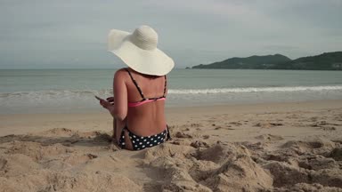Kumsalda güneşlenme sırasında cep telefonu kullanan genç kadın. Bikini ve güneş şapkalı güzel kız resimleri alır ve kısa mesaj yazıyor