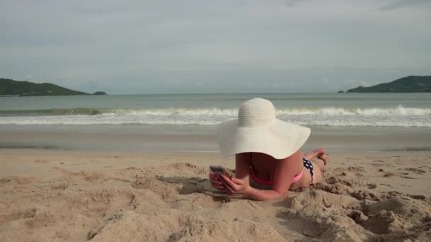 Jovem usando celular enquanto toma sol na praia. Menina bonita em um biquíni e chapéu de sol tira fotos e escreve mensagem de texto — Vídeo de Stock