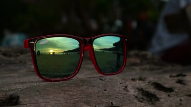 Gafas de sol acostadas en la arena al atardecer. En unas gafas de sol reflejadas mar, olas y cielo . — Vídeos de Stock