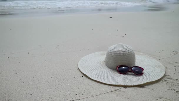 Sunglasses and white broad-brim lying on a sand. seascape with waves on sandy shore. — Stock Video