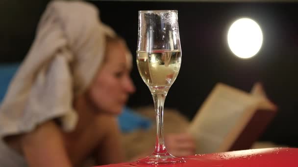 Lo champagne primo piano scorre in un bicchiere. Bella donna sdraiata su un letto e legge un libro. rallentatore — Video Stock