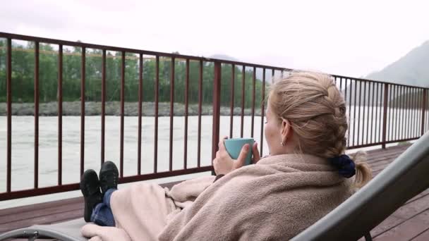 Młoda kobieta pije gorącą kawę lub herbatę w pobliżu górskiej rzeki. Spokojny i przytulny czas w mountain resort. — Wideo stockowe