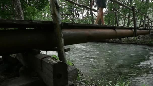 Jeune femme marche le long d'un étroit pont en bois sur une rivière de montagne. camping et concept d'aventure — Video