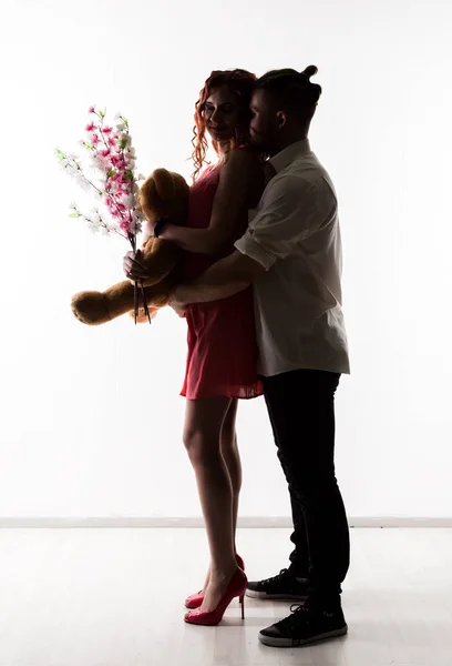 Chica con un chico en una cita. Hombre sosteniendo flores y mujer sosteniendo oso de peluche. silueta sobre un fondo claro —  Fotos de Stock