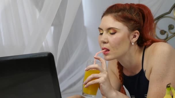 Молодая женщина наслаждается на кровати, используя ноутбук и пьет апельсиновый сок. замедленное движение — стоковое видео