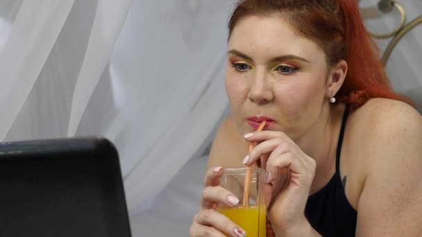 Mujer joven disfrutando en una cama, utilizando el ordenador portátil y bebe jugo de naranja. cámara lenta — Vídeos de Stock