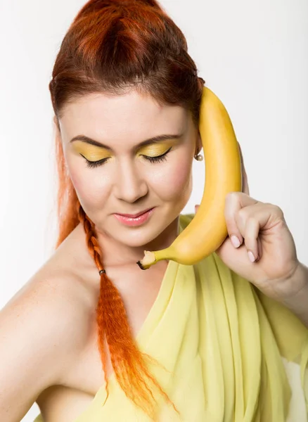 Jonge roodharige vrouw met banaan in de buurt van haar gezicht als smartphone op een witte achtergrond. — Stockfoto