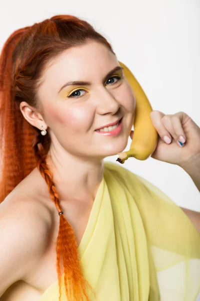 Jonge roodharige vrouw met banaan in de buurt van haar gezicht als smartphone op een witte achtergrond. — Stockfoto