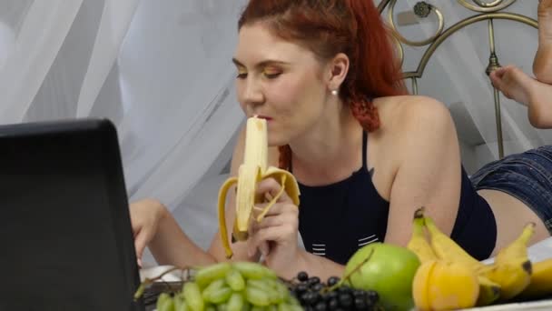 Νεαρή γυναίκα απολαμβάνοντας σε ένα κρεβάτι, χρησιμοποιώντας φορητό υπολογιστή και τρώει μια μπανάνα στο σπίτι το πρωί. αργή κίνηση — Αρχείο Βίντεο
