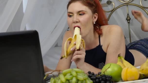 Jonge vrouw genieten op een bed, het gebruik van de laptop en het eten van een banaan thuis in de ochtend. Slow motion — Stockvideo