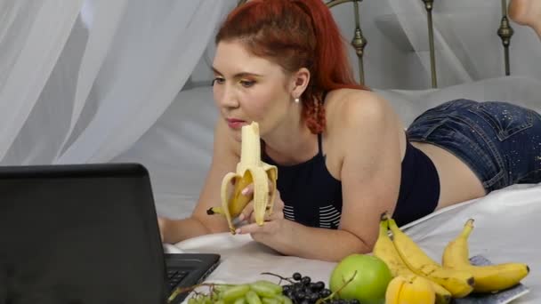 Mujer joven disfrutando en una cama, utilizando el ordenador portátil y comer un plátano en casa por la mañana. cámara lenta — Vídeos de Stock