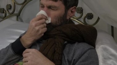 Hasta bir adam bir yatak sıcak bir eşarp ile onun boynuna bir soğuk veya grip sırasında burun akıntısı sahip olarak onun burun üfleme yatıyor. 4k