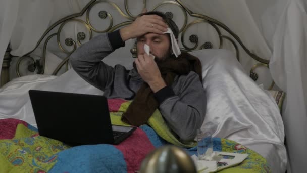 Chory leży w łóżku z ciepłym szalikiem wokół jego szyi i ręcznik na jego głowę, wideo rozmowy z lekarzem na laptopie. zwolnionym tempie — Wideo stockowe