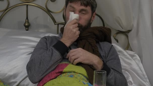 A sick man lies in a bed with a warm scarf around his neck, blowing his nose as having a runny nose during a cold or influenza. 4K — Stock Video