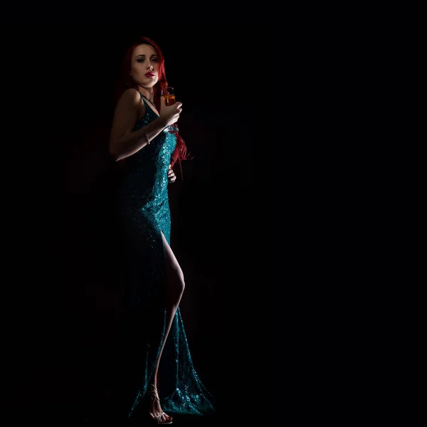 Giovane rossa donna sexy in abito elegante blu in possesso di bicchieri da vino con champagne e posa in un buio. spazio libero per il testo — Foto Stock