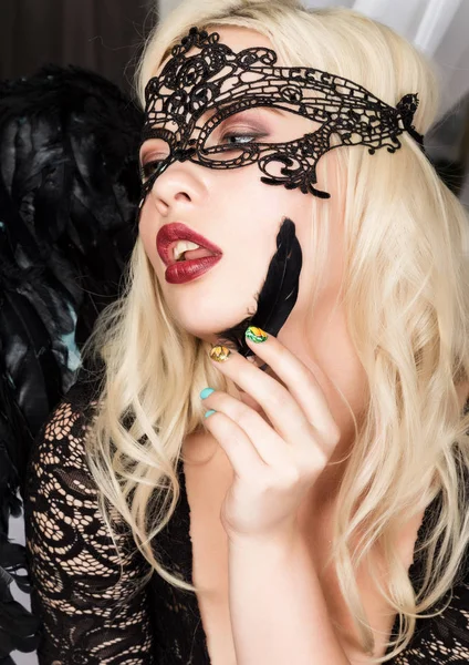 Sexy mujer rubia en una máscara de encaje sosteniendo una pluma negra cerca de su cara — Foto de Stock