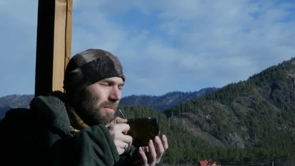 Одинокий бородатый мужчина, стоящий на смотровой площадке и смотрящий на гору и пьющий кофе или чай из большой чашки. замедленное движение — стоковое видео