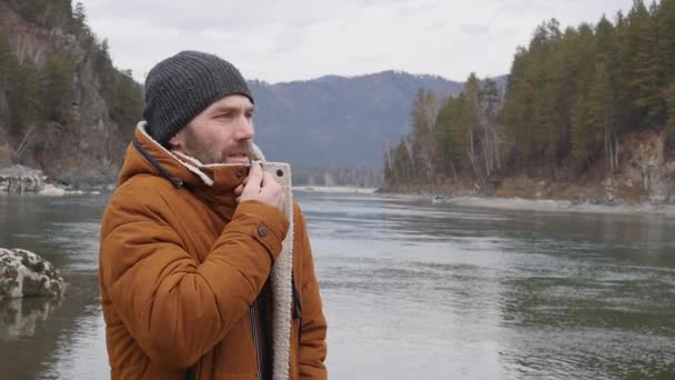 Joven hombre guapo se encuentra en la orilla del río de montaña en tiempo frío. cámara lenta — Vídeos de Stock
