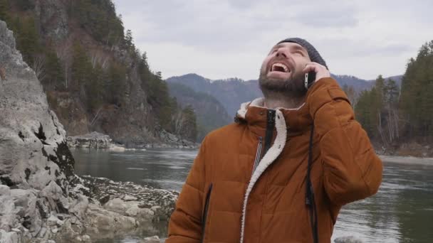 Joven hombre guapo está en la orilla del río de montaña en tiempo frío, tipo barbudo hablando por teléfono. cámara lenta — Vídeos de Stock