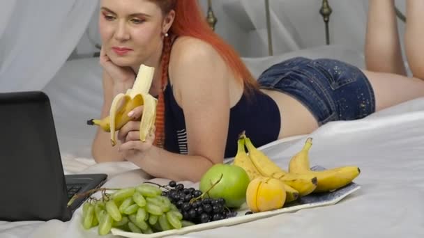 Young woman enjoying on a bed, using laptop and eating a banana at home in the morning. slow motion — Stock Video