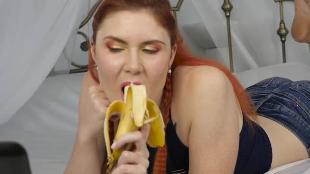 Νεαρή γυναίκα απολαμβάνοντας σε ένα κρεβάτι, χρησιμοποιώντας φορητό υπολογιστή και τρώει μια μπανάνα στο σπίτι το πρωί. αργή κίνηση — Αρχείο Βίντεο