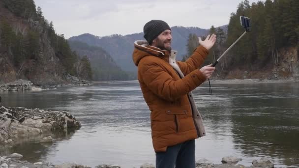 Fotografías turísticas barbudas a sí mismo en un teléfono móvil en la orilla de un río de montaña. cámara lenta — Vídeo de stock