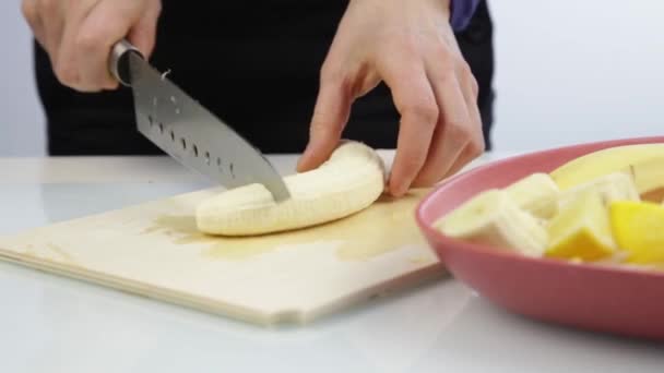 As mãos das mulheres cortam uma banana em uma placa de cozinha de madeira com uma faca — Vídeo de Stock