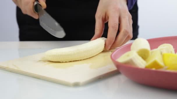 Frauenhände schneiden mit einem Messer eine Banane auf einem hölzernen Küchenbrett — Stockvideo