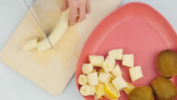 Las manos femeninas cortan el plátano en la tabla de madera de la cocina. Vista superior. 4K — Vídeo de stock