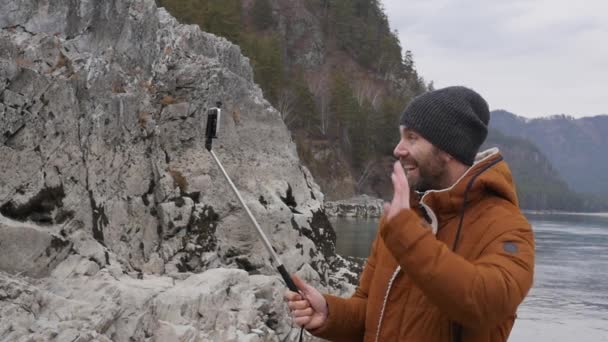 Бородатий турист проводить відеоконференцію з друзями на мобільному телефоні на березі гірської річки. повільний рух — стокове відео