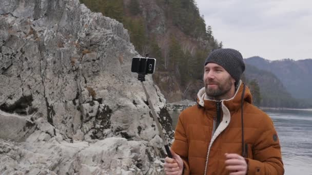 Turista barbudo está transmitiendo vídeo en un teléfono móvil en la orilla de un río de montaña. cámara lenta — Vídeos de Stock