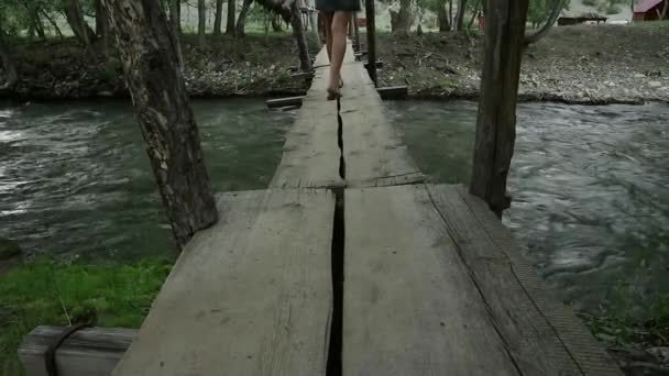 Fiatal nő egy hegyi folyó mentén keskeny fahíd gyaloglás. kemping és kaland fogalma — Stock videók