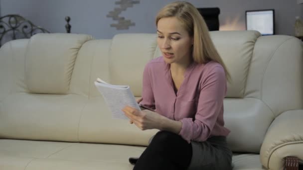 Mulher loira em um sofá lê suas notas em um caderno e verifica notas com o smartphone — Vídeo de Stock
