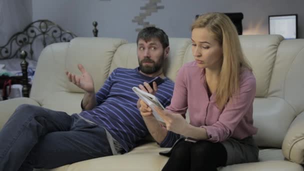 Блондинка на дивані читає нотатки в блокноті, а її чоловік сидить поруч і спілкується по телефону — стокове відео