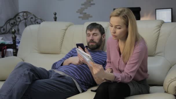 Estudiante rubia lee sus notas, y su novio sentado en el sofá y utiliza teléfonos inteligentes — Vídeos de Stock