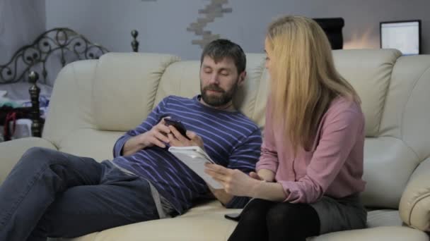 Blonde Studentin liest ihre Notizen, ihr Freund sitzt auf dem Sofa und nutzt Smartphones — Stockvideo