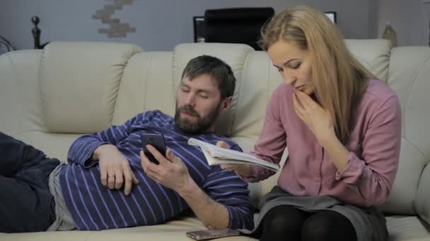 Studente biondo legge i suoi appunti, e il suo ragazzo seduto sul divano e utilizza smartphone — Video Stock