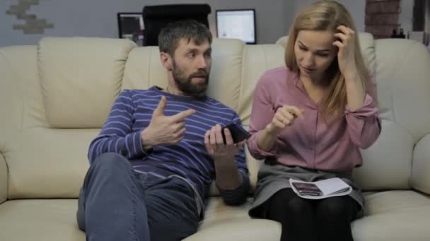 Blonde vrouw op een sofa leest haar notities in een notitieblok en haar man in de buurt zitten en chatten op de telefoon — Stockvideo
