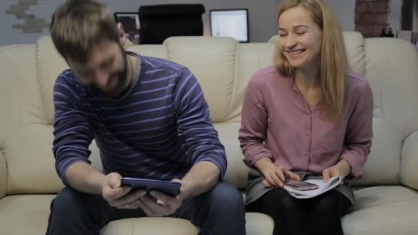 Блондинка читает свои заметки, а ее парень сидит на диване и пользуется смартфонами — стоковое видео