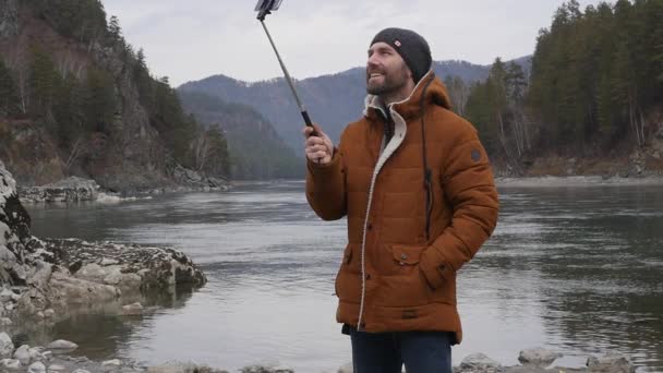 Turista barbudo está transmitiendo vídeo en un teléfono móvil en la orilla de un río de montaña. cámara lenta — Vídeos de Stock
