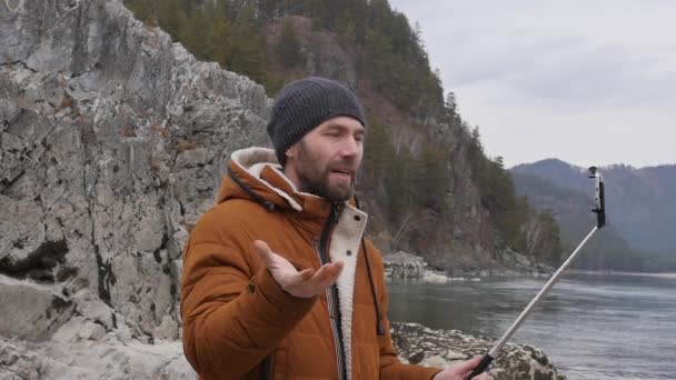 Turista barbuto sta avendo videoconferenza con gli amici su un telefono cellulare sulla riva di un fiume di montagna. rallentatore — Video Stock