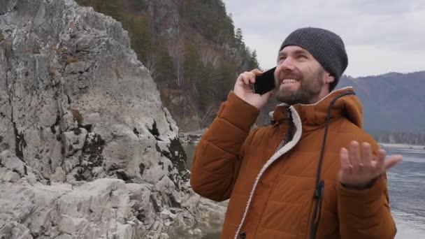 Turista barbudo está teniendo videoconferencia con amigos en un teléfono móvil en la orilla de un río de montaña. cámara lenta — Vídeos de Stock