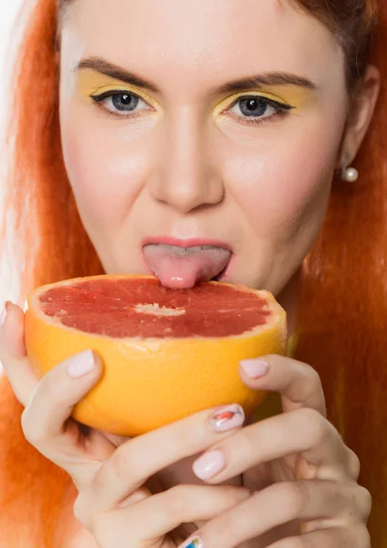 Jonge roodharige vrouw likt een grapefruit op een witte achtergrond. begrip van gezond eten — Stockfoto