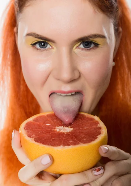 Unga rödhårig kvinna slickar en grapefrukt på en vit bakgrund. begreppet hälsosam kost — Stockfoto